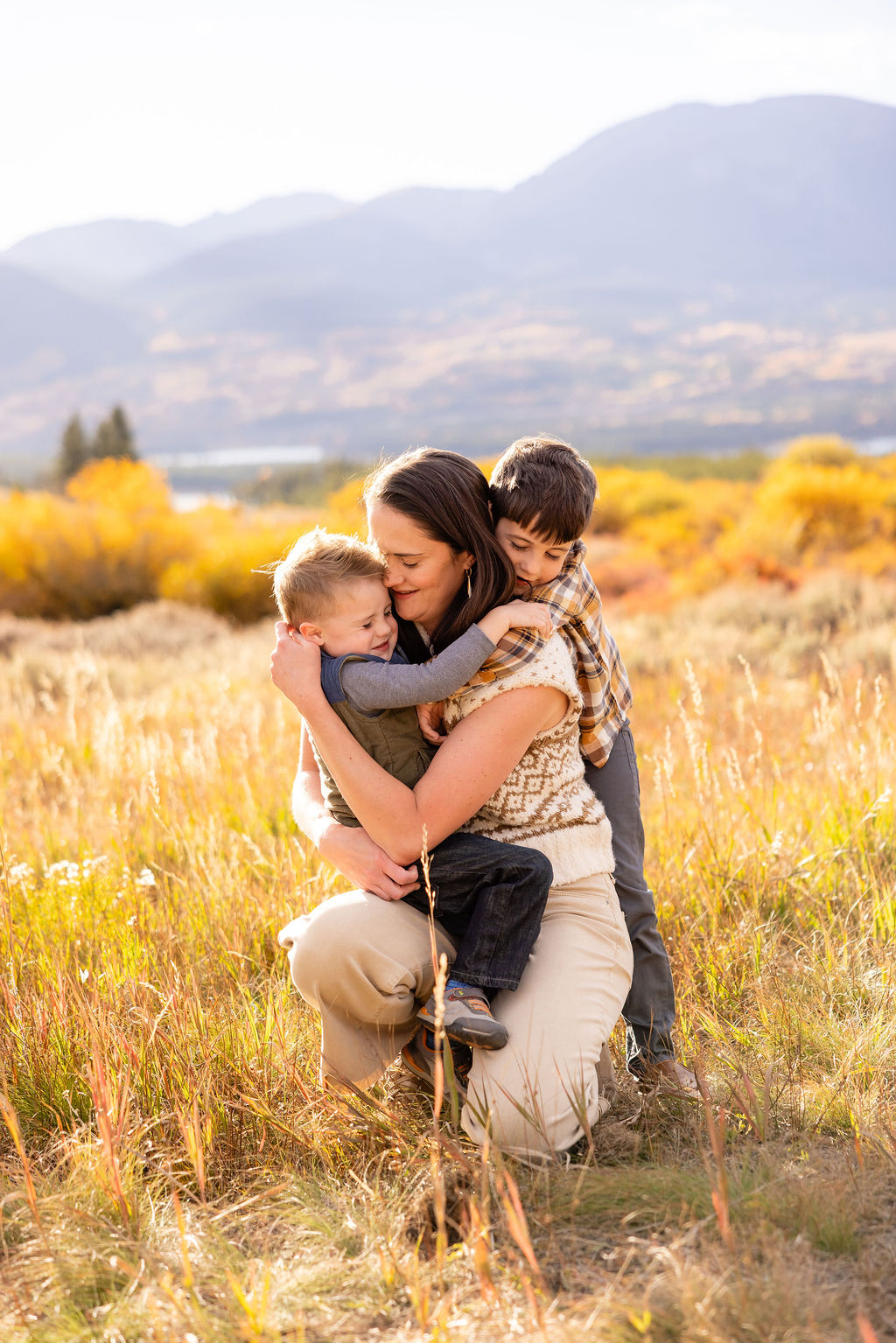 A mother kneeling in a sunlit fall meadow embracing her two young children